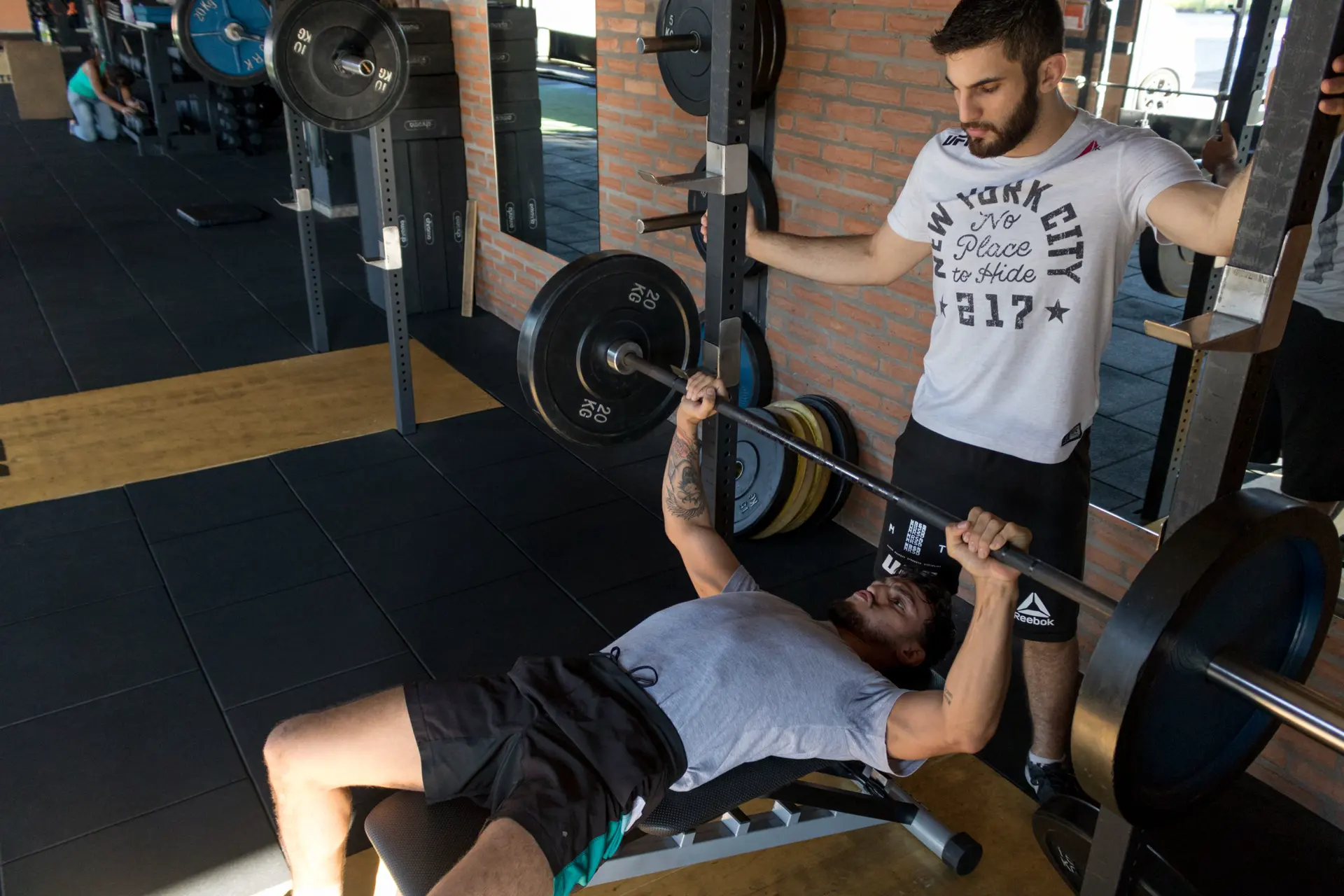 Powerlifter bench presses a heavy barbell while a spotter guards the bar in a strength gym